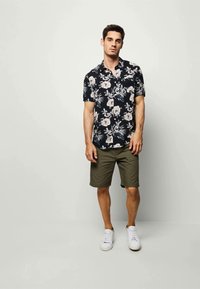 Chemise à manches courtes en tissu noir avec des motifs floraux blancs, accompagnée de shorts vert olive et de baskets blanches, se tenant devant un mur clair.