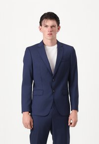 Marineblaues tailliertes Sakko mit Reverskragen, zwei Knöpfen und seitlichen Taschen. Getragen über einem weißen Hemd, glatte Stoffstruktur.