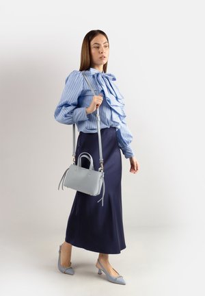 Donna con camicetta a righe blu e gonna blu scuro, che porta una borsa grigio chiaro e scarpe slingback con tacco basso abbinato, in piedi davanti a uno sfondo bianco.