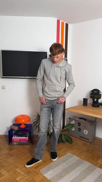 Sweatshirt cinza com um bolso frontal e detalhe de botão, combinado com jeans cinza claro e sapatos pretos sem atacadores; o ambiente interior possui uma decoração colorida.