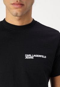 Černé bavlněné tričko s vroubkovaným kulatým výstřihem. Má bílý potisk loga "KARL LAGERFELD JEANS" umístěný na levé straně.