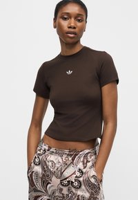 Νέα γυναίκα με εφαρμοστό σκούρο καφέ κοντομάνικο crop top με μικρό λευκό λογότυπο Adidas και παντελόνια με σχέδια, με τα χέρια στις τσέπες.