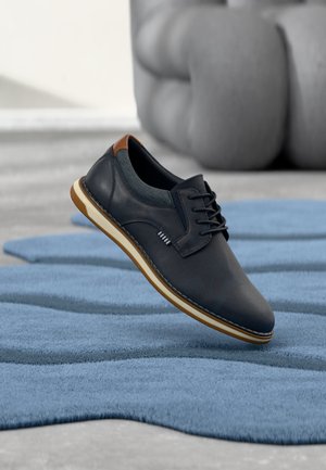 Chaussure décontractée noire à lacets avec détail talon marron flottant au-dessus d'un tapis bleu texturé, avec une chaise grise rembourrée en arrière-plan.