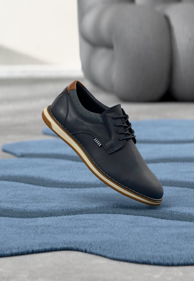 Chaussure décontractée noire à lacets avec détail talon marron flottant au-dessus d'un tapis bleu texturé, avec une chaise grise rembourrée en arrière-plan.