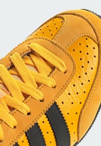 Gelbe Leder-Sneaker mit schwarzen Streifen, perforierten Details und flachen gelben Schnürsenkeln. Texturierte Oberfläche mit matter Optik.