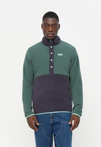Fleece pullover in donker groen en marineblauw met hoge kraag, knoopsluiting en lichte aqua accenten. Beschikt over een logo op de borst.