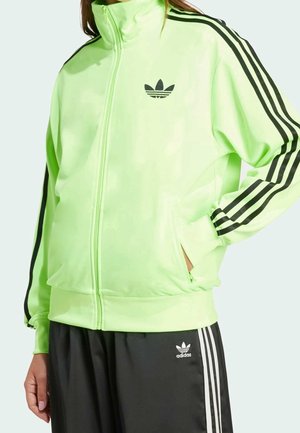 Person iført en neongrøn Adidas træningsjakke med sorte striber på ærmerne og sorte Adidas træningsbukser med hvide striber.