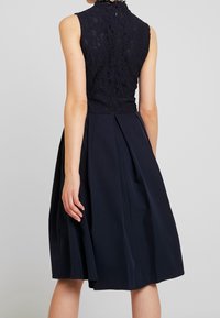 Marinblå klänning med spetsliv, hög halsringning och ärmlös design, samt en plisserad kjol av mjukt tyg.