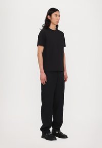 Svart t-skjorte med korte ermer og løse svarte bukser. Modellen står oppreist, iført svarte sko med sølvdetaljer. Minimalistisk design.