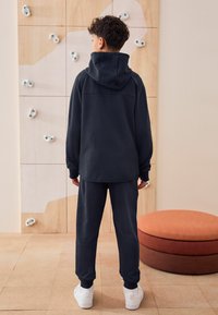 Donkerblauwe hoodie en bijpassende joggingbroek met ribbelmanchetten, gecombineerd met witte sneakers. Achtergrond bevat een klimwand en een rond oranje kussen.
