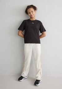 Čierne bavlnené tričko s bielym logom, v kombinácii so svetlobežovými teplákmi so zipsami po stranách a čiernymi športovými topánkami s bielymi podrážkami.