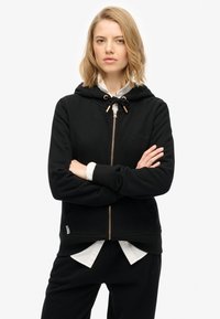 Il hoodie nero con zip presenta un cappuccio regolabile, polsini a coste e un logo ricamato. Indossato sopra una camicia bianca con colletto. Tessuto morbido.