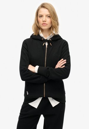 Il hoodie nero con zip presenta un cappuccio regolabile, polsini a coste e un logo ricamato. Indossato sopra una camicia bianca con colletto. Tessuto morbido.