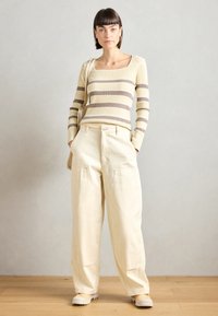 Pulover cu dungi și nervuri în galben și maro, asortat cu pantaloni lejeri de culoare crem. Pantofii de culoare deschisă completează ținuta.
