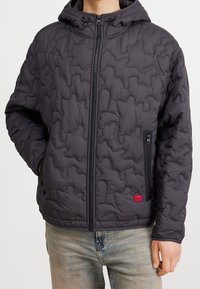 Dunkelgraue gepolsterte Kapuzenjacke mit gestepptem Muster, Vorderreißverschluss und seitlichen Taschen; mit einem kleinen roten Logotag.