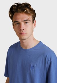Blaues Baumwoll-T-Shirt mit lockerer Passform, Rundhalsausschnitt und einem gestickten Hasenmotiv auf der linken Brust. Weiche Textur, minimalistische Details.