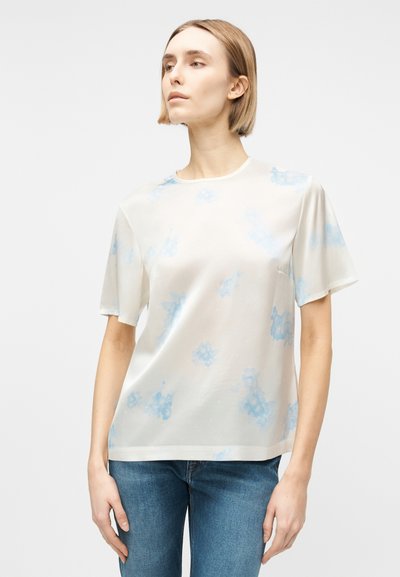 Γυναίκα με κοντά ξανθά μαλλιά που φοράει ένα άνετο λευκό μπλουζάκι με ανοιχτά μπλε αφηρημένα σχέδια και μπλε τζιν, στέκεται μπροστά σε ένα απλό φόντο.