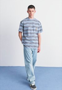 Blåstripet t-skjorte med lomme, kombinert med lyseblå baggy jeans. Modellen har på seg svarte og hvite sneakers. Ensfarget bakgrunn.
