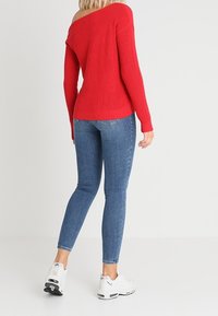 Červený úpletem ribbed svetřík bez ramenního rukávu v kombinaci s modrými úzkými džíny. Bílé tenisky dotváří celý outfit. Důraz na texturu a přiléhavý střih.
