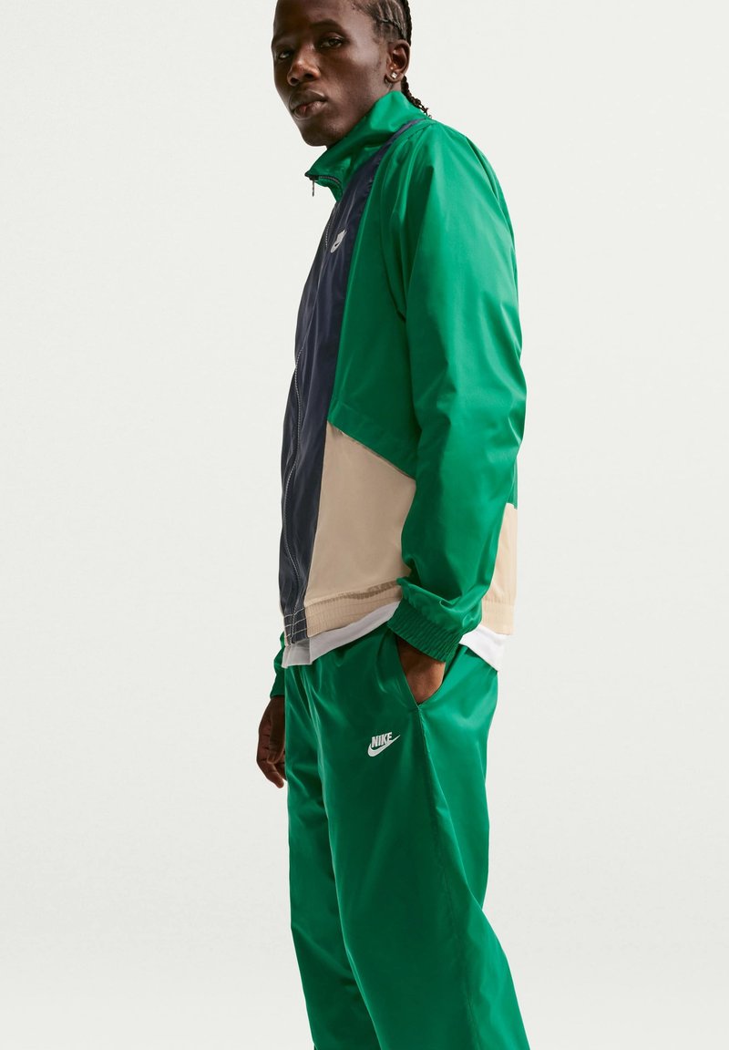 Person trägt eine grüne, marineblaue und beige Nike-Jacke mit passenden grünen Nike-Hosen und steht mit einer Hand in der Tasche vor einem einfarbigen Hintergrund.