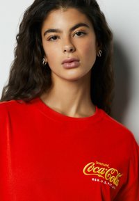 Czerwona bawełniana koszulka z dużym żółtym haftowanym logo Coca-Cola na przodzie. Krótkie rękawy i okrągły dekolt.