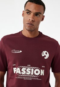 Bordeaux kortemouw t-shirt met witte geprinte graphics met de tekst "Doel," "PASSIE," en "PSM." Soepele stof met ronde hals.