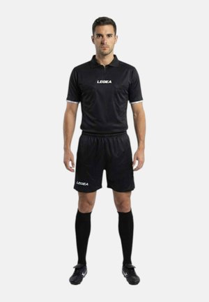 Uomo in piedi che indossa una divisa nera da arbitro composta da maglietta, pantaloncini, calzini al ginocchio e scarpe da calcio su sfondo bianco neutro.