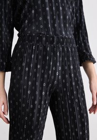Combinaison noire plissée dotée d'une taille cintrée, de manches longues et d'un motif de petits diamants réfléchissants sur l'ensemble.