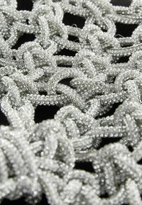 Sidabrinės karoliukų apdailos su sudėtingais mazgų raštais, turinčiais tekstūrinį paviršių ir atspindinčius akcentus, sluoksniuotos tamsiame fone.