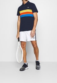 Tmavě modré polo tričko se žlutými, červenými, oranžovými a modrými vodorovnými pruhy, v kombinaci s bílými šortkami s logem, držící tenisovou raketu.