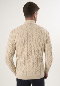 Beige kabelstrikket sweater med ribbet kant og manchetter, der har teksturerede mønstre og en kravehalset. Vist fra bagsiden.