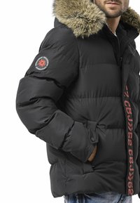 Schwarze Steppjacke mit pelzgefütterter Kapuze, vorne mit einem auffälligen roten vertikalen Logo und einem runden Patch am linken Ärmel.