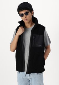 Zwarte fleece vest met een ritssluiting aan de voorkant, twee voorzakken en een borstzak van contrasterende stof. Gecombineerd met een grijze T-shirt en een zonnebril.