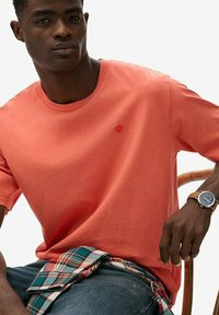 Koralfarvet bomuld T-shirt med rund halsudskæring og korte ærmer, med en lille logo. Båret over denim, fremhævet med en ternet skjorte bundet om livet.