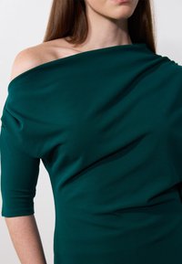 Sjøgrønn off-shoulder kjole laget av mykt stoff, med drapert detaljer og trekvartlange ermer. Glatt tekstur med subtile folder.