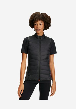 Frau mit lockigem Haar, die eine ärmellose schwarze gesteppte Weste über einem schwarzen Kurzarmshirt und schwarzen Hosen trägt, stehend vor einem weißen Hintergrund.