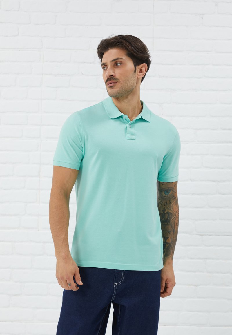 Polo shirt turquoise clair à manches courtes avec une patte de boutonnage à trois boutons, porté avec un jean bleu foncé. Présente une texture lisse.