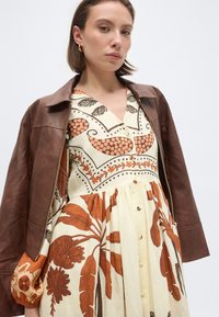 Γυναίκα που φοράει καφέ δερμάτινο μπουφάν πάνω από ένα κρεμ φόρεμα με πορτοκαλί βοτανικά και γεωμετρικά σχέδια, στέκεται μπροστά σε ένα απλό ανοιχτό χρωματικό φόντο.