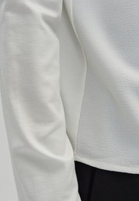 Top bianco a maniche lunghe con superficie testurizzata, con una sezione in tessuto liscio sul lato. L'orlo si trova sopra i pantaloni neri.