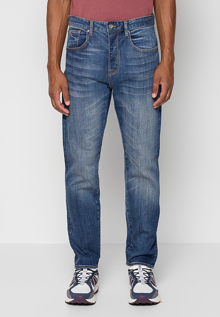 Uomo che indossa jeans blu sbiaditi, sneaker bianche e blu navy, e una camicia bordeaux, in piedi contro uno sfondo chiaro e semplice.