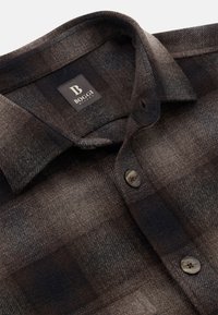 Camisa de franela a cuadros en tonos marrón oscuro y gris, con cuello clásico, cierre de botones y una textura tejida. Etiqueta de la marca visible.