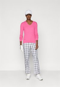 Roosa ribadega V-kaelusega sviiter, millele on sobitatud must-valged ruudulised püksid; mudel kannab valget mütsi ja golfikindaid, seistes sportlikes kingades.