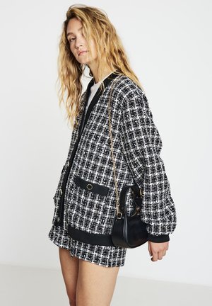 Veste en tweed noir et blanc avec bords côtelés et deux poches, associée à une mini-jupe noire et un petit sac à main noir avec une bandoulière chaîne.
