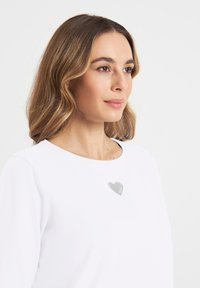 Witte langefsleevetop met een ronde hals en een zilveren hartaccent op de borst, gemaakt van een gladde stof.