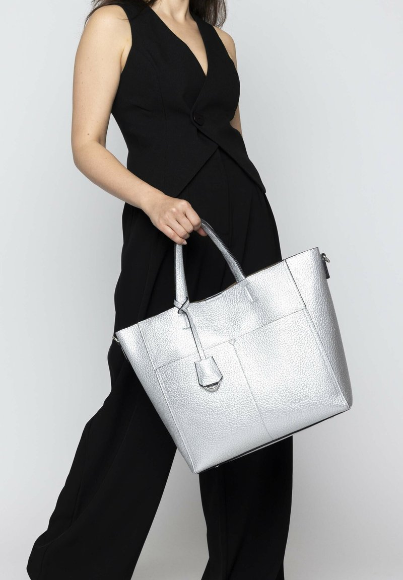 Silver leather tote bag with a textured finish, two handles, and front pocket detail, held by a person in a black outfit.