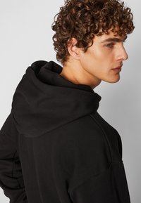 Sudadera negra con capucha de doble capa y corte relajado. La tela parece suave, con detalles de costura visibles en los hombros.