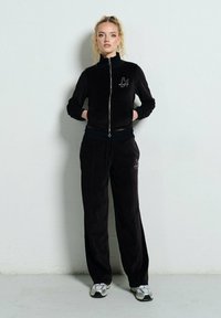 Kobieta stoi przy ścianie, ubrana w czarny dres z weluru, składający się z kurtki na zamek i spodni o szerokich nogawkach, w połączeniu z srebrnymi trampkami.