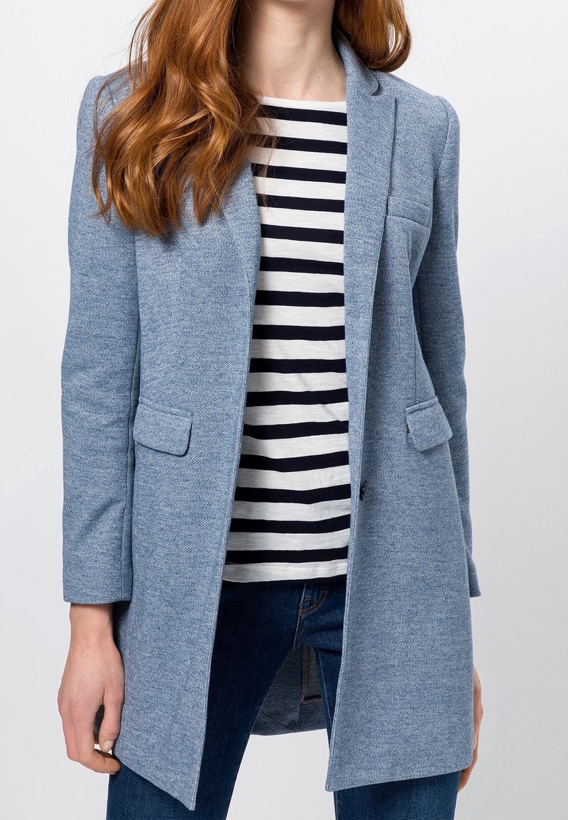 Femme portant un blazer bleu clair par-dessus une chemise blanche à rayures noires avec un jean bleu foncé, debout devant un fond uni.