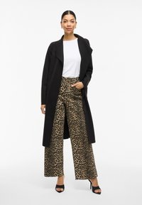 Cappotto lungo nero sopra una maglietta bianca, abbinato a pantaloni larghi con stampa leopardata. Sandali con tacco quadra scuri esaltano l'aspetto audace dell'outfit.