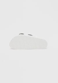 White footwear with a ridged sole. Features two adjustable straps with metallic accents. Smooth, minimalist design without patterns.